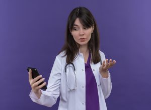 confused young female doctor in medical robe with stethoscope looks at phone and holds hand straight on isolated violet background with copy space